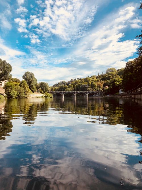 Free Peaceful river scene with a historic bridge under a vibrant blue sky and lush trees. Stock Photo