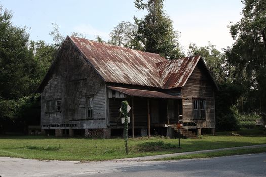 An old, weathered house with a rusty roof stands in an overgrown area surrounded by trees.