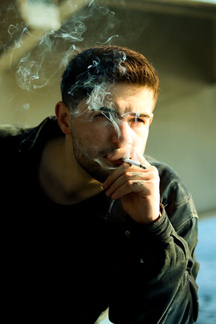 A thoughtful man in a denim jacket smoking a cigarette, enveloped in smoke in a contemplative pose.