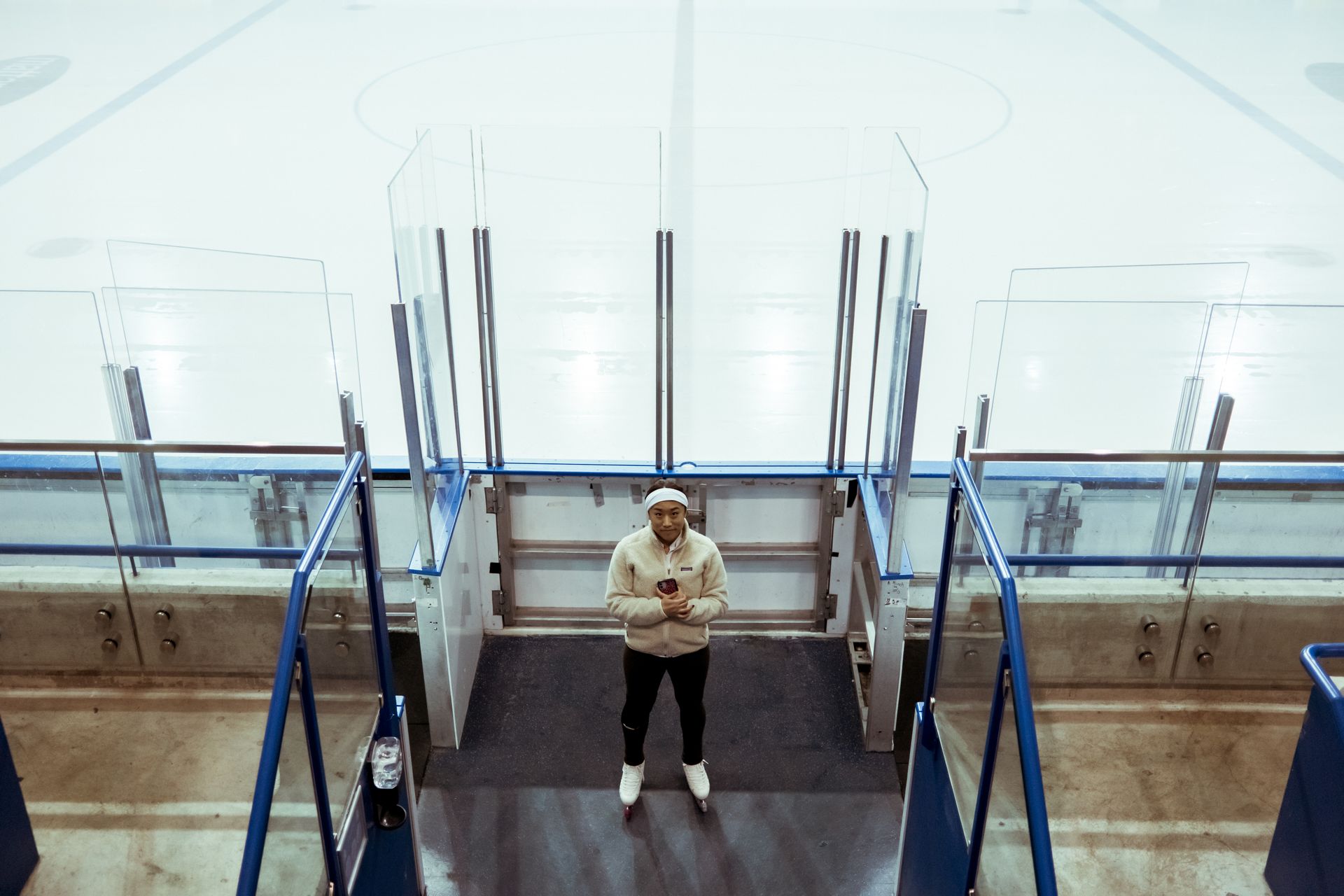 An Asian woman standing at the entrance of an ice skating rink wearing sportswear.