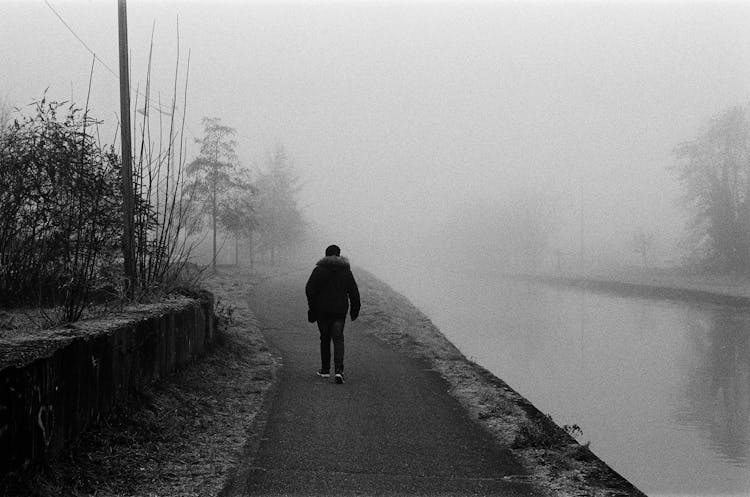 A Grayscale Photo Of A Person In Black Jacket Walking Near The Lake