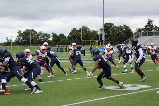 Football players in action during a competitive match on a green field under cloudy skies.