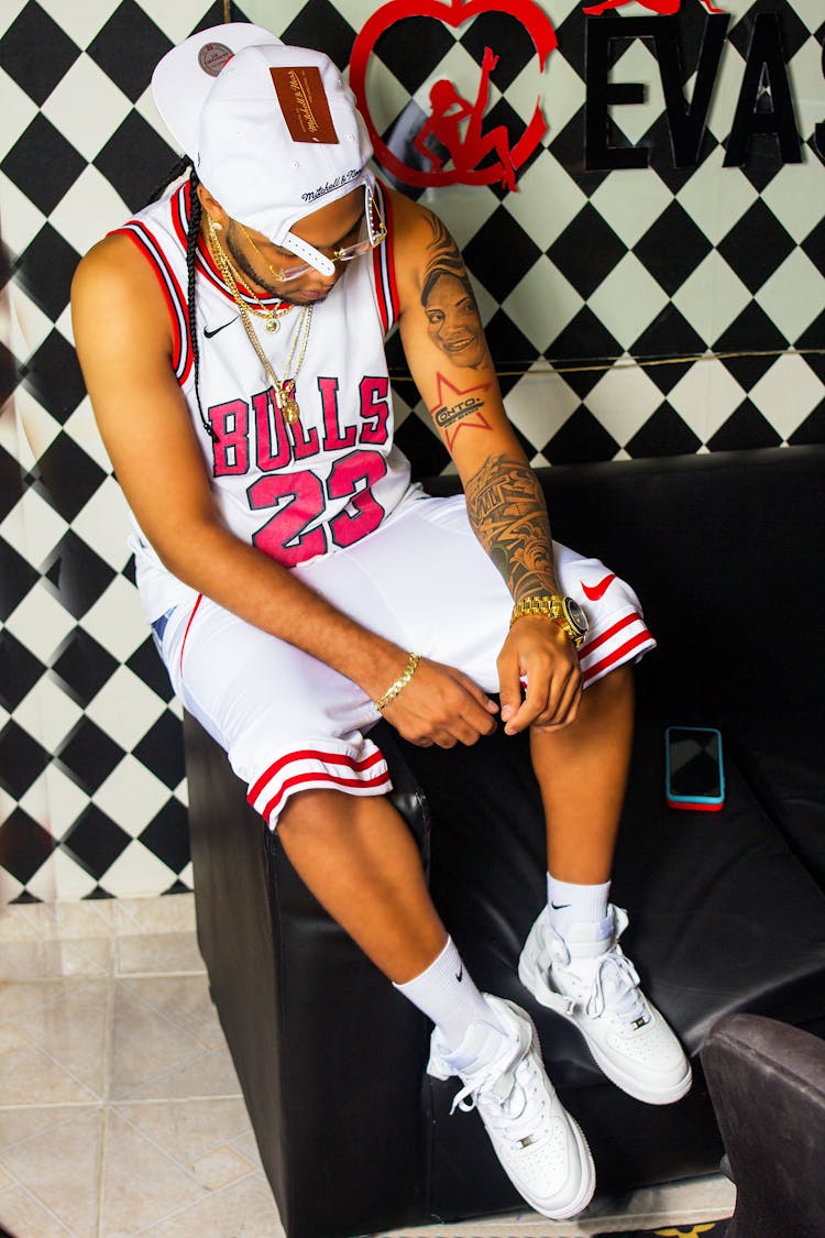 A Tattooed Man In A Basketball Jersey Sitting On The Armrest Of A Couch