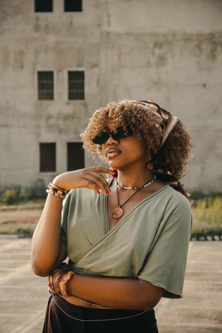 Portrait of a fashionable woman wearing sunglasses and jewelry, smiling outdoors on a sunny day in an urban setting.