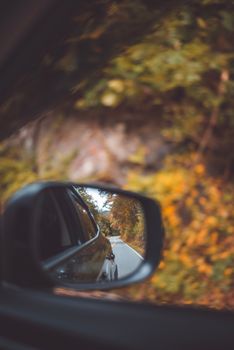 Close-up of car side mirror reflecting a scenic autumn road with blurred foliage.