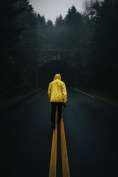 A solitary figure in a yellow jacket walks down a foggy road towards a dark tunnel.