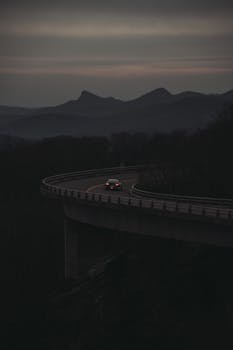 A car travels along a winding highway with mountains in the background at twilight.