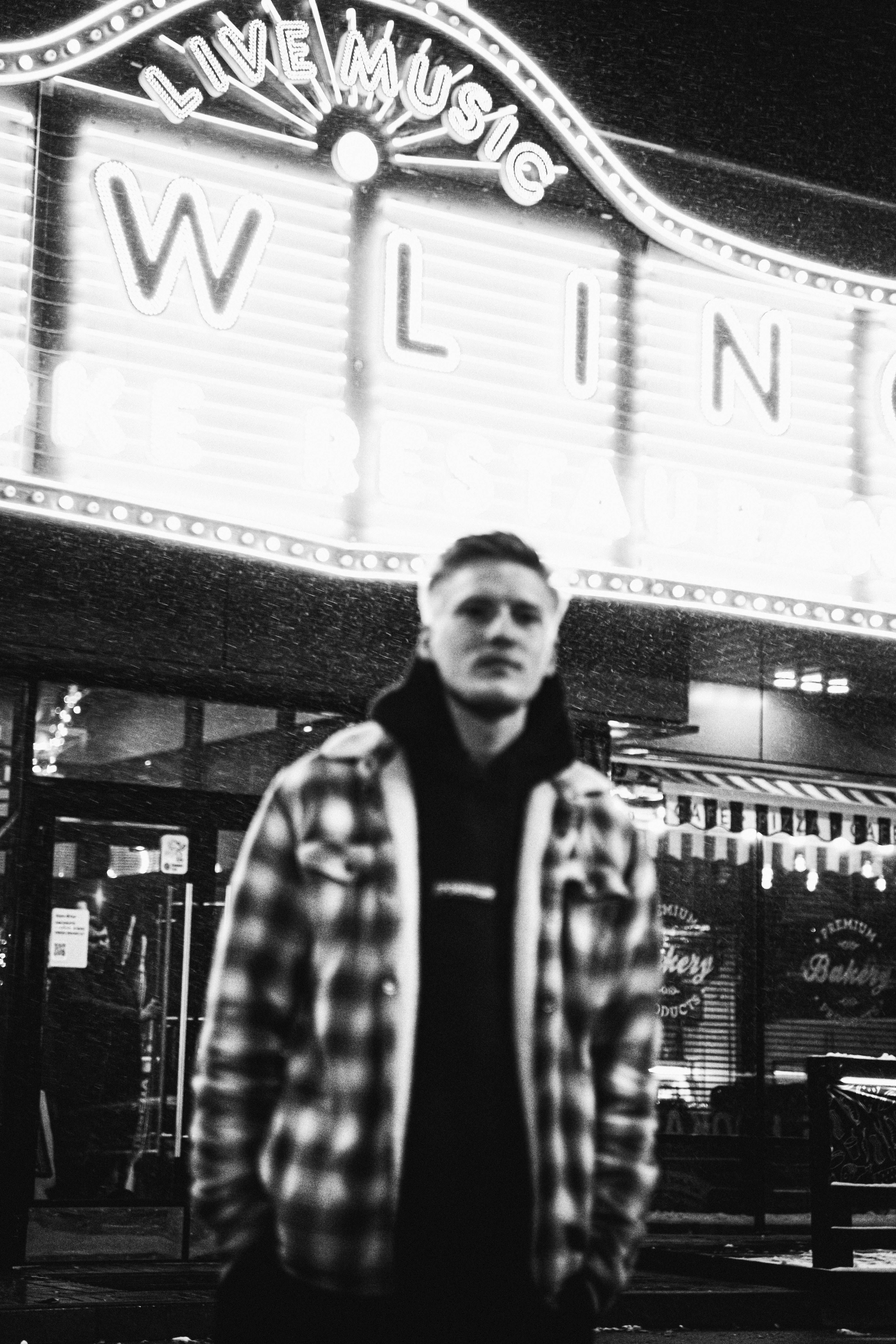 Black and white photo of a man in plaid jacket with neon signage background.
