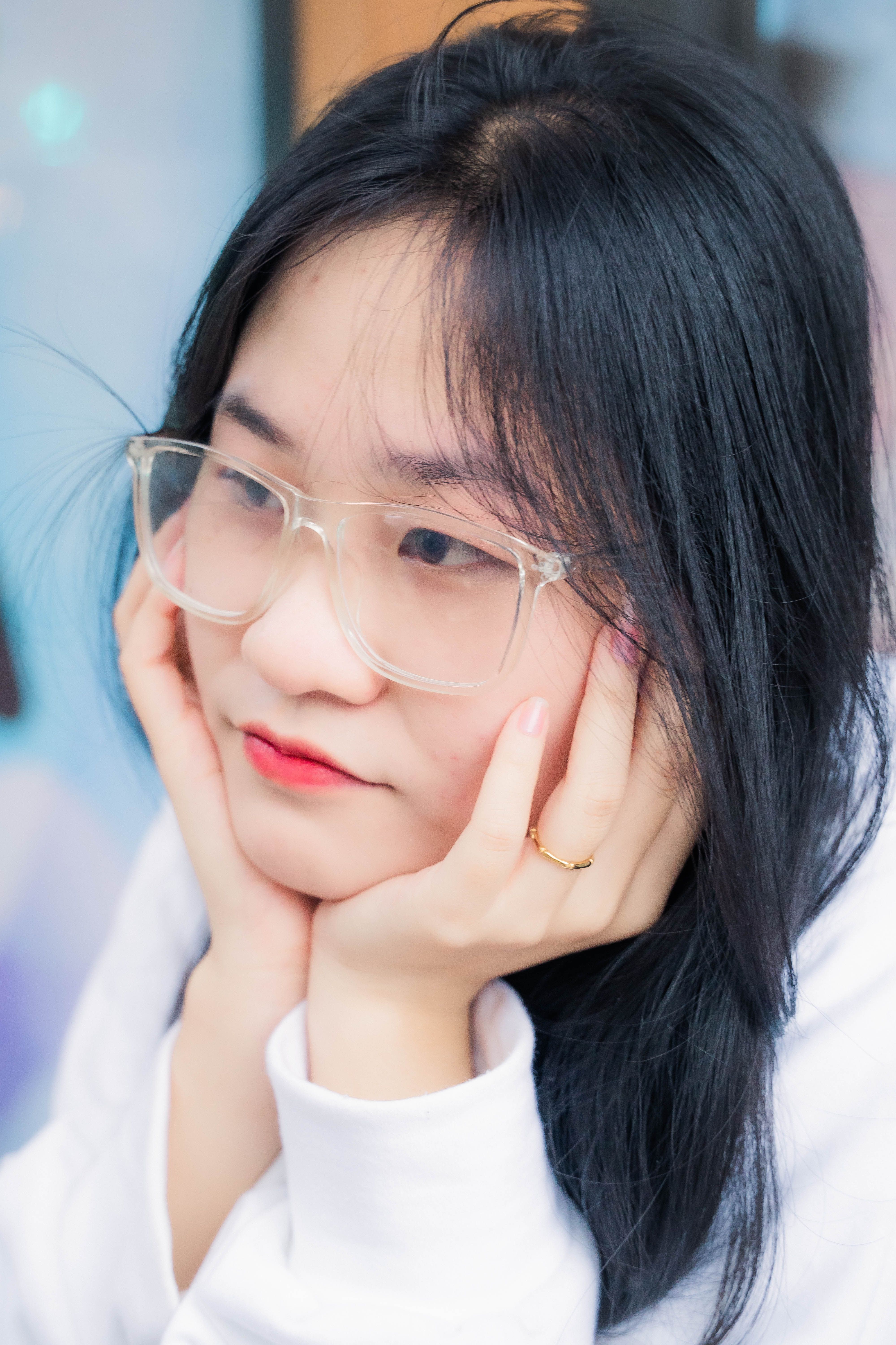 Portrait of a young Asian woman with clear glasses, resting her chin on her hands.