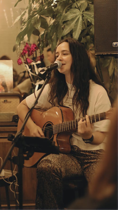 A Woman in White Shirt Playing Guitar while Singing on Microphone