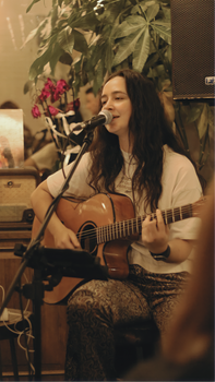 A woman in a white shirt sings and plays guitar in a cozy indoor setting, creating a warm and inviting atmosphere.