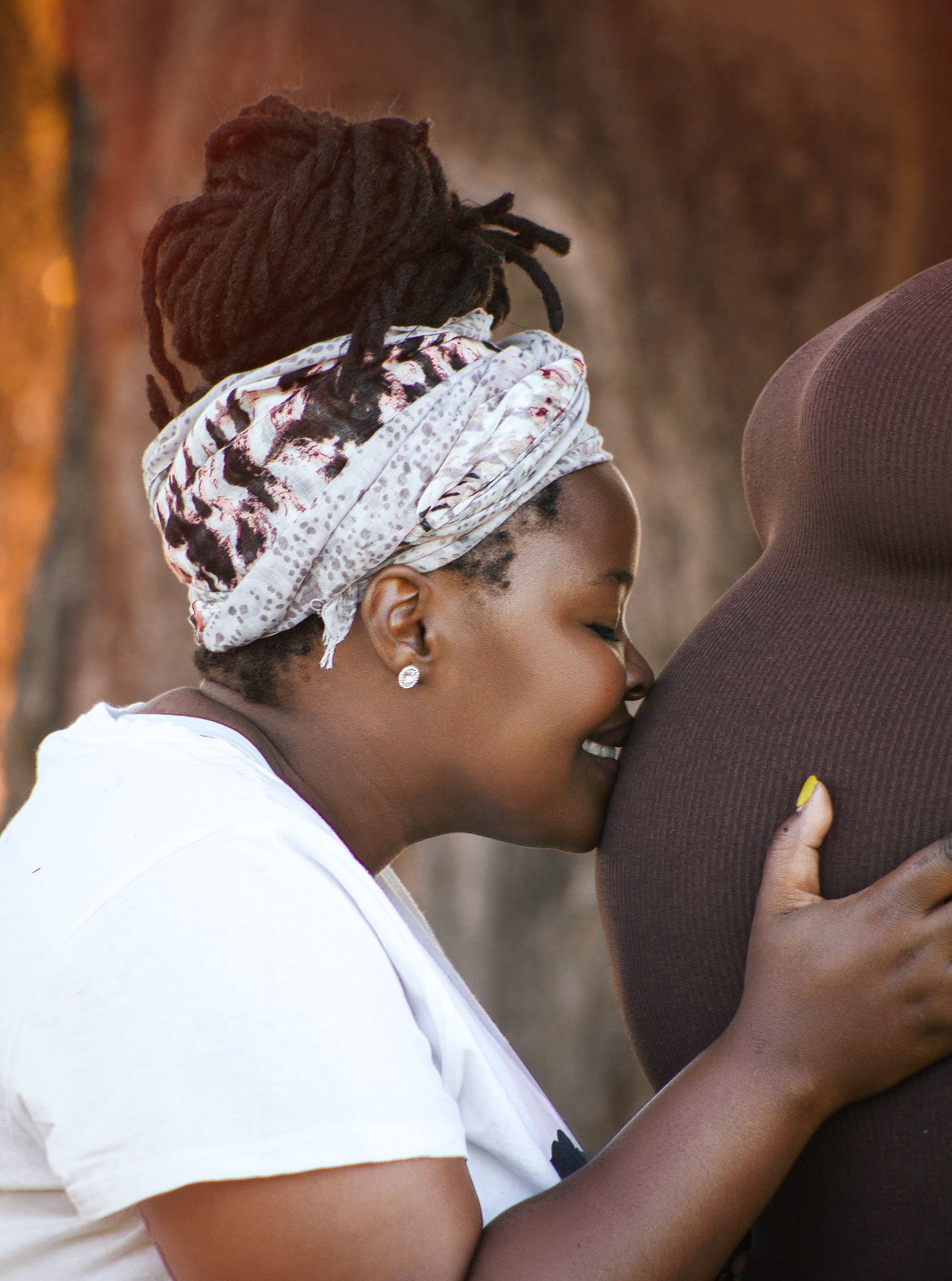 Two Black women outdoors in warm daylight; one in a white shirt and headwrap leans her face against the pregnant belly of the other, hand resting gently, both calm and engaged.