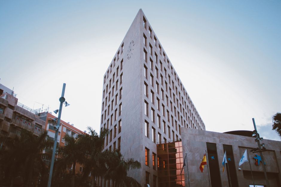 Una toma arquitectónica de un edificio gubernamental moderno en una ciudad con banderas visibles y un reloj al anochecer.