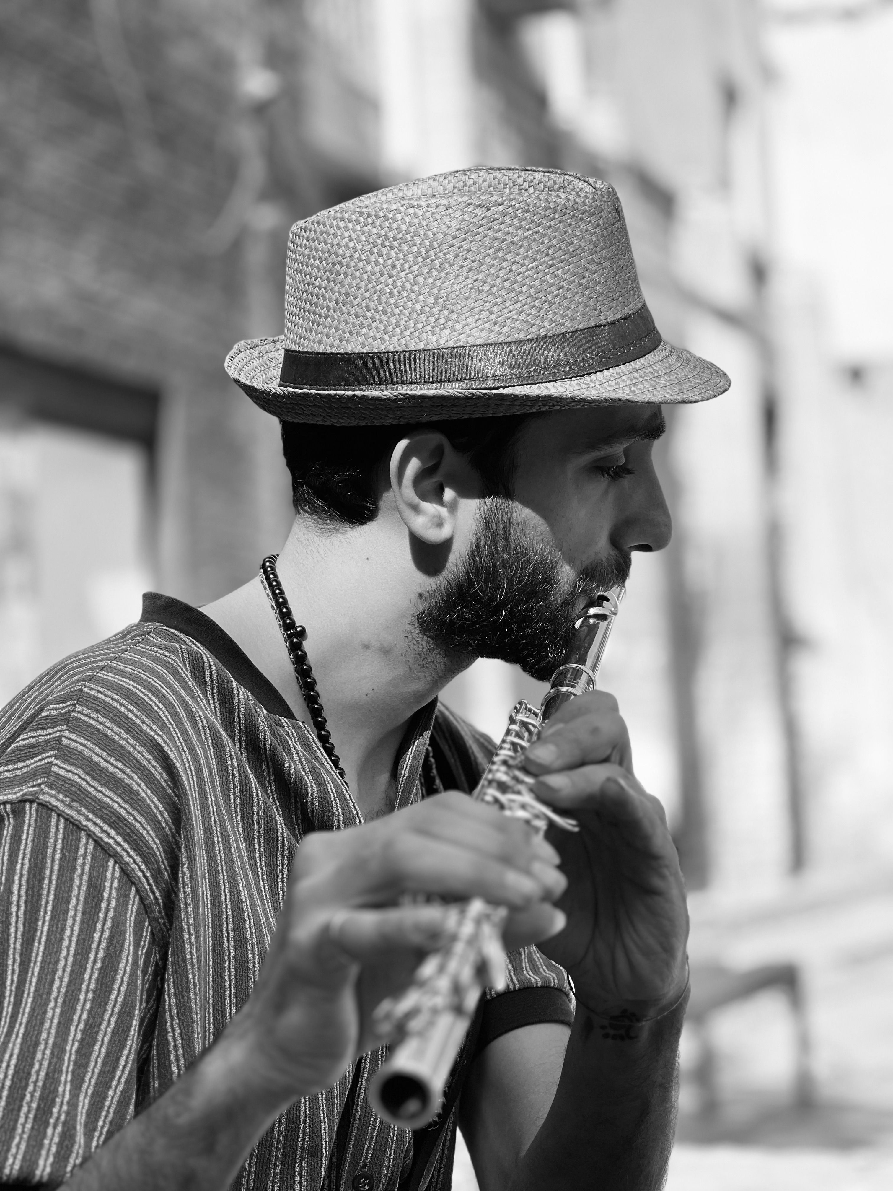 Black and white portrait of a bearded musician playing flute outdoors, wearing a straw hat and striped shirt.