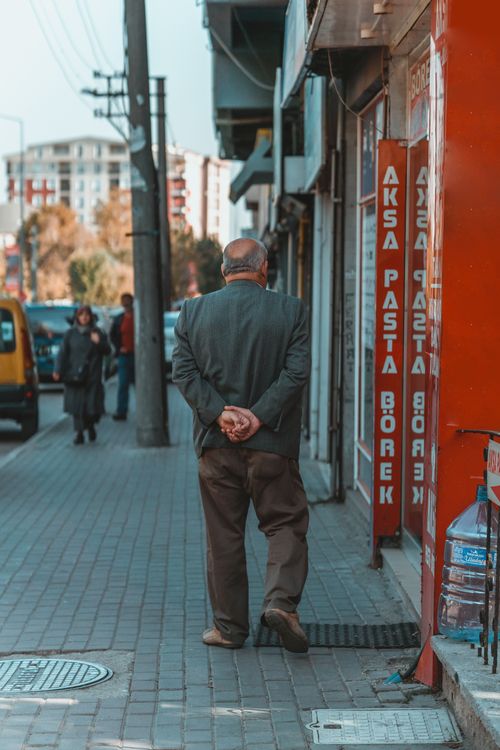 Free An elderly man walking on a city sidewalk, surrounded by storefronts and urban scenery. Stock Photo