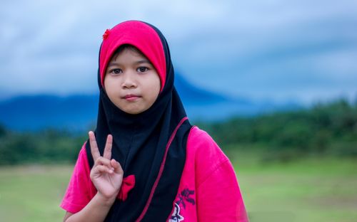 Free Portrait of a young girl wearing a hijab, making a peace sign, set against a serene outdoor background. Stock Photo