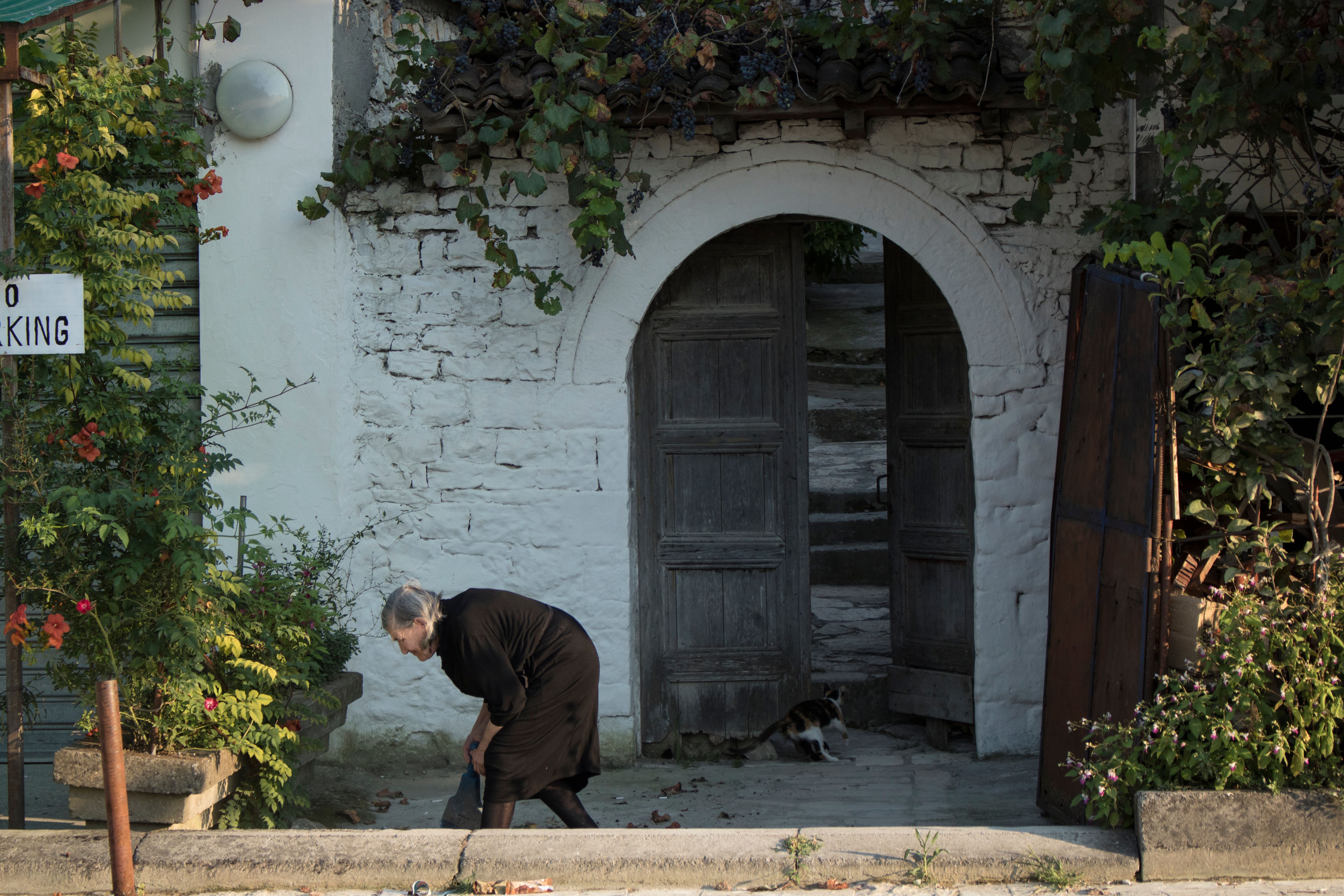 Elderly woman bends near rustic doorway in quiet street setting.