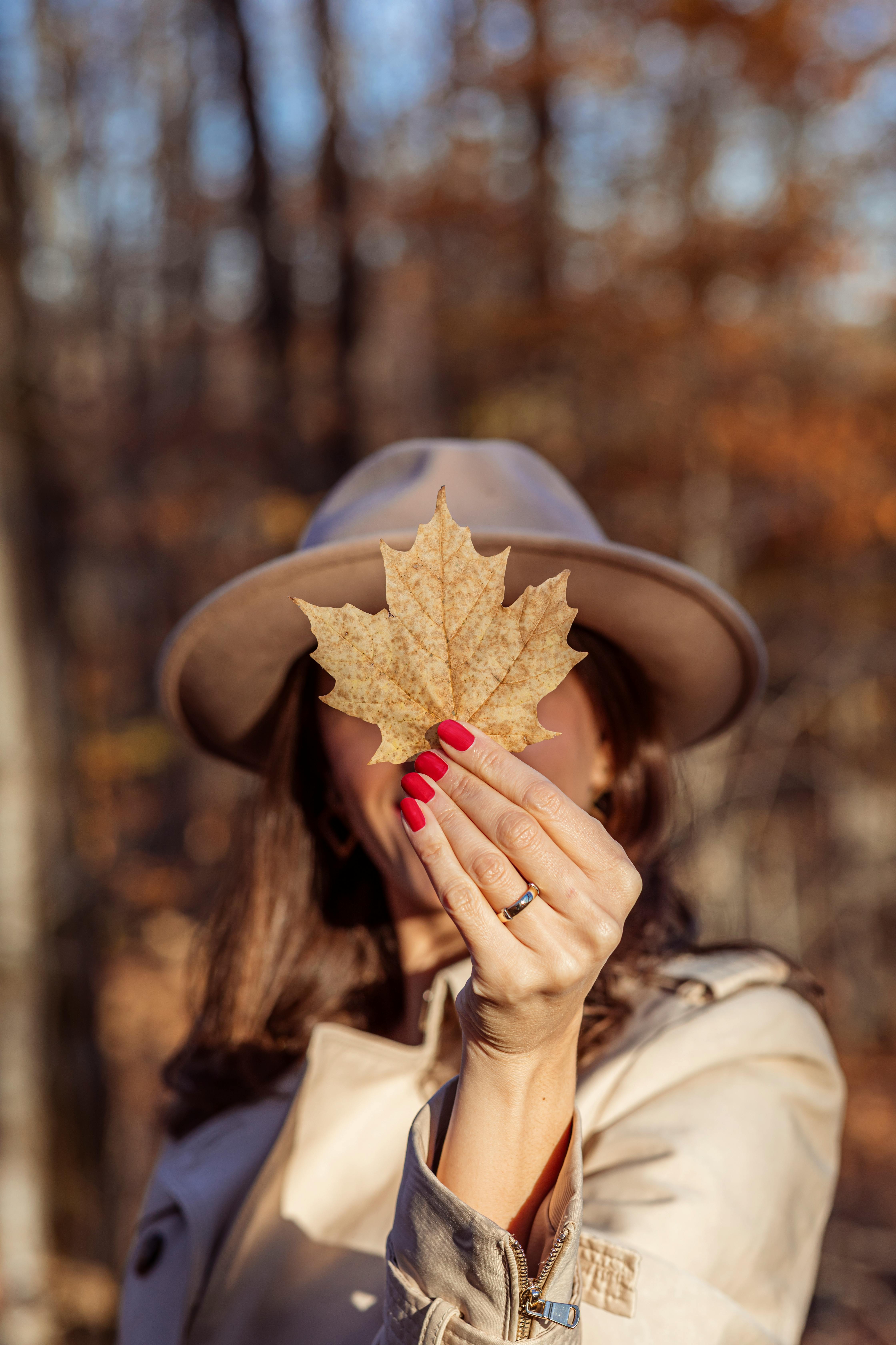 Close-up of a woman in a hat holding a dried maple leaf in a fall forest.