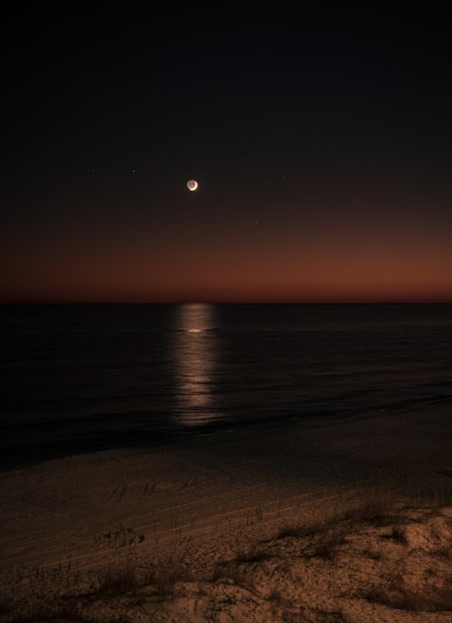 Free A serene night scene of the moon casting its reflection on the ocean surface, viewed from a sandy beach. Stock Photo