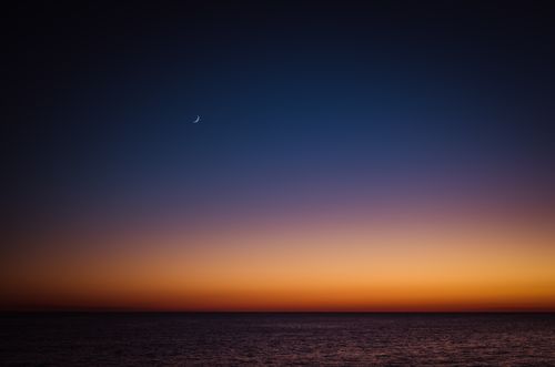 Free A stunning sunset over the ocean with a crescent moon in the sky, creating a dramatic and peaceful scene. Stock Photo