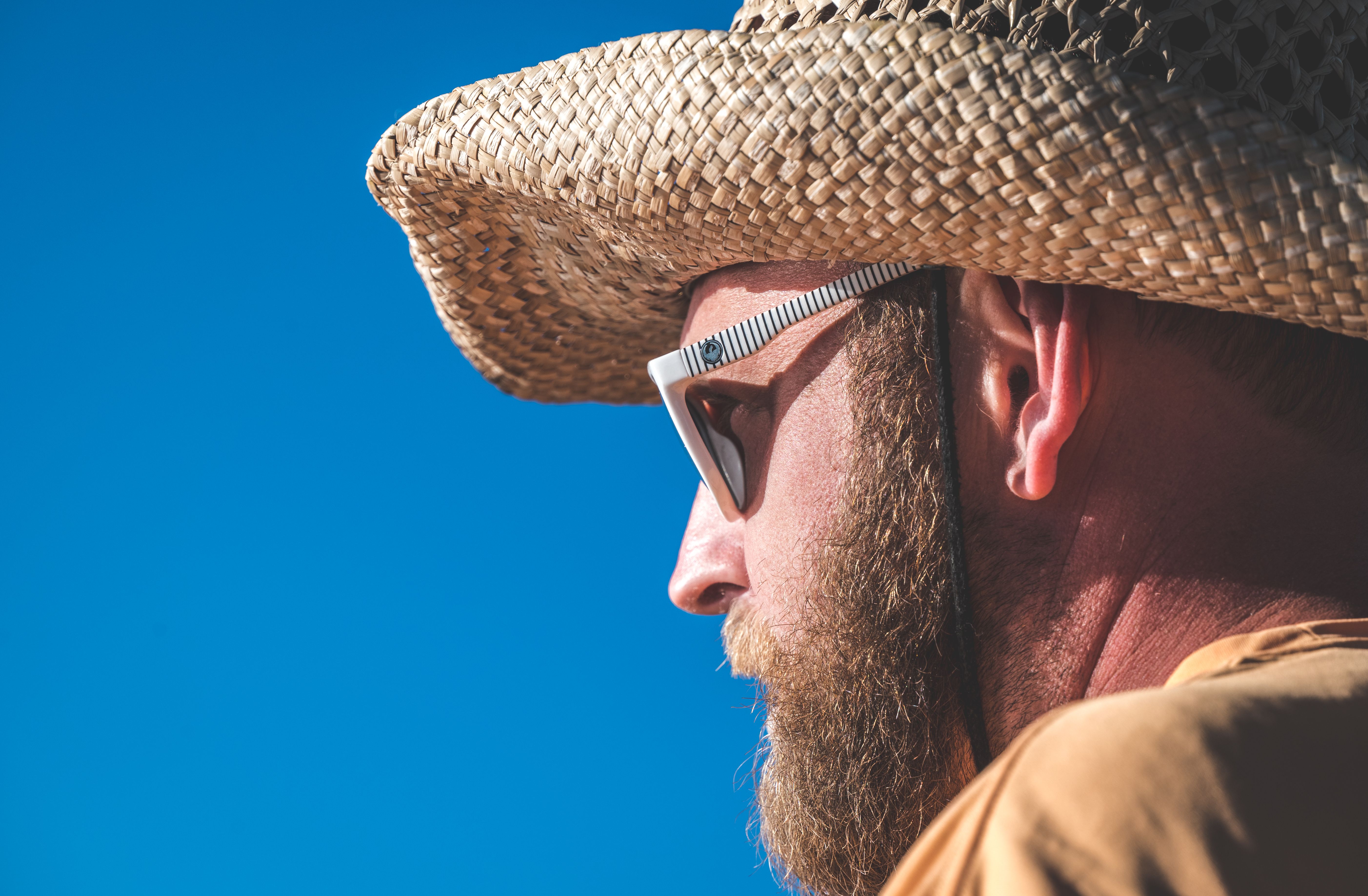 Bearded man wearing a straw hat and sunglasses under clear blue sky, enjoying a sunny day.