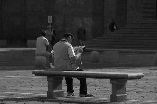 Free Black and white photo of a senior man reading a newspaper on a stone bench in a plaza. Stock Photo