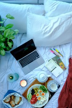 Flat lay of a cozy bedroom scene featuring a laptop, breakfast tray, and candles on a bed.