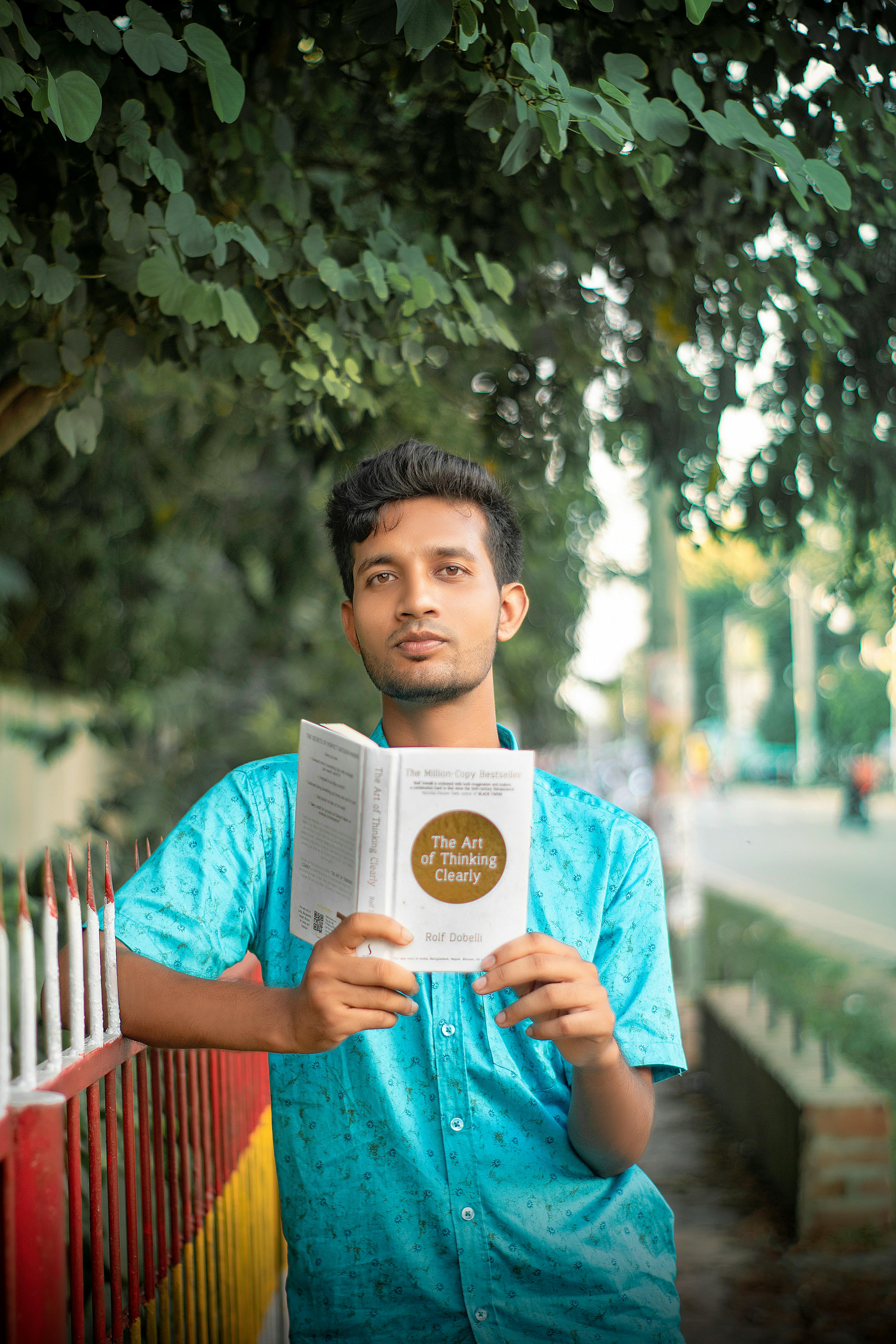 A young adult reads a book while standing on a street lined with trees on a sunny day.