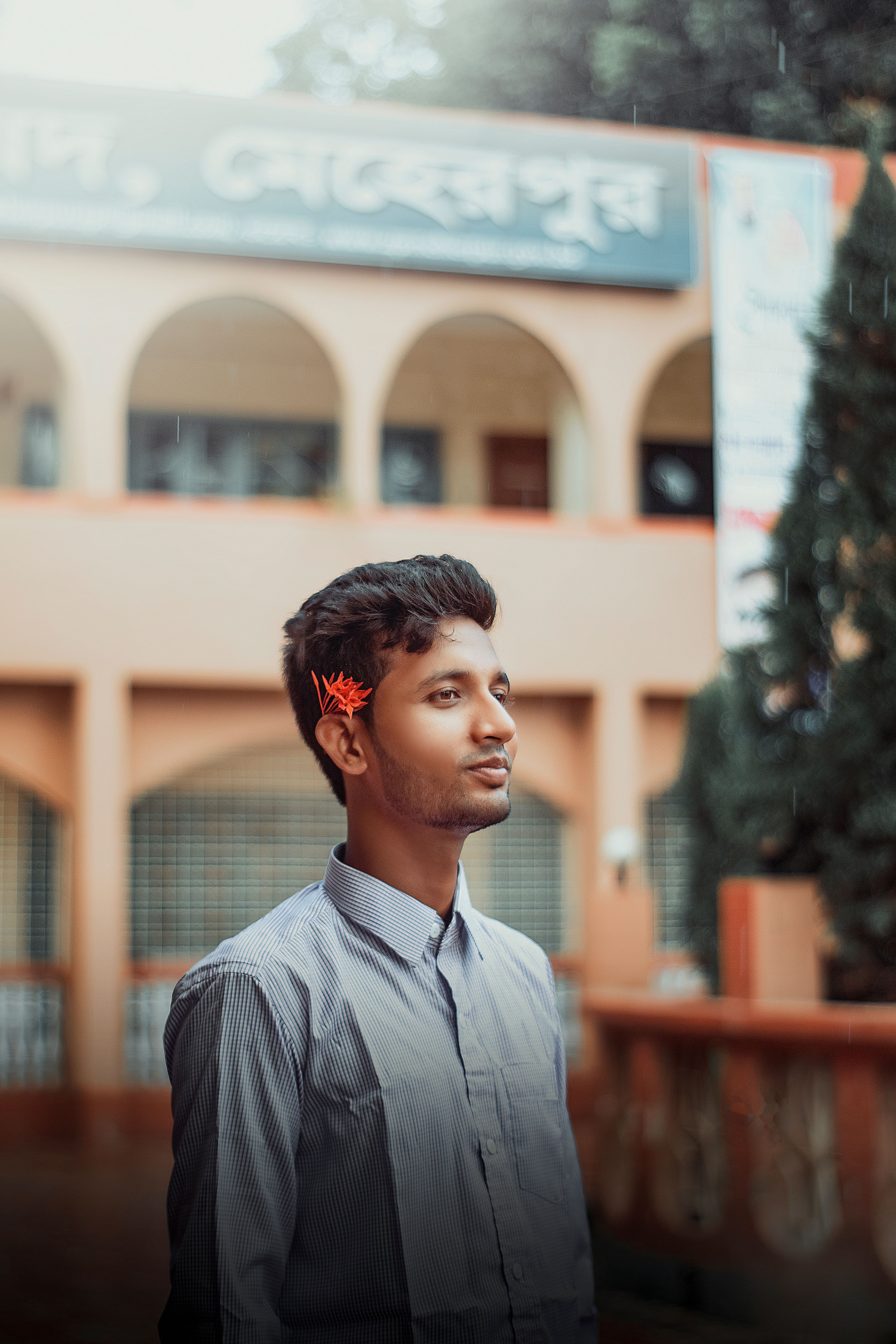 Asian man with a flower in his hair, smiling outdoors with architectural background.