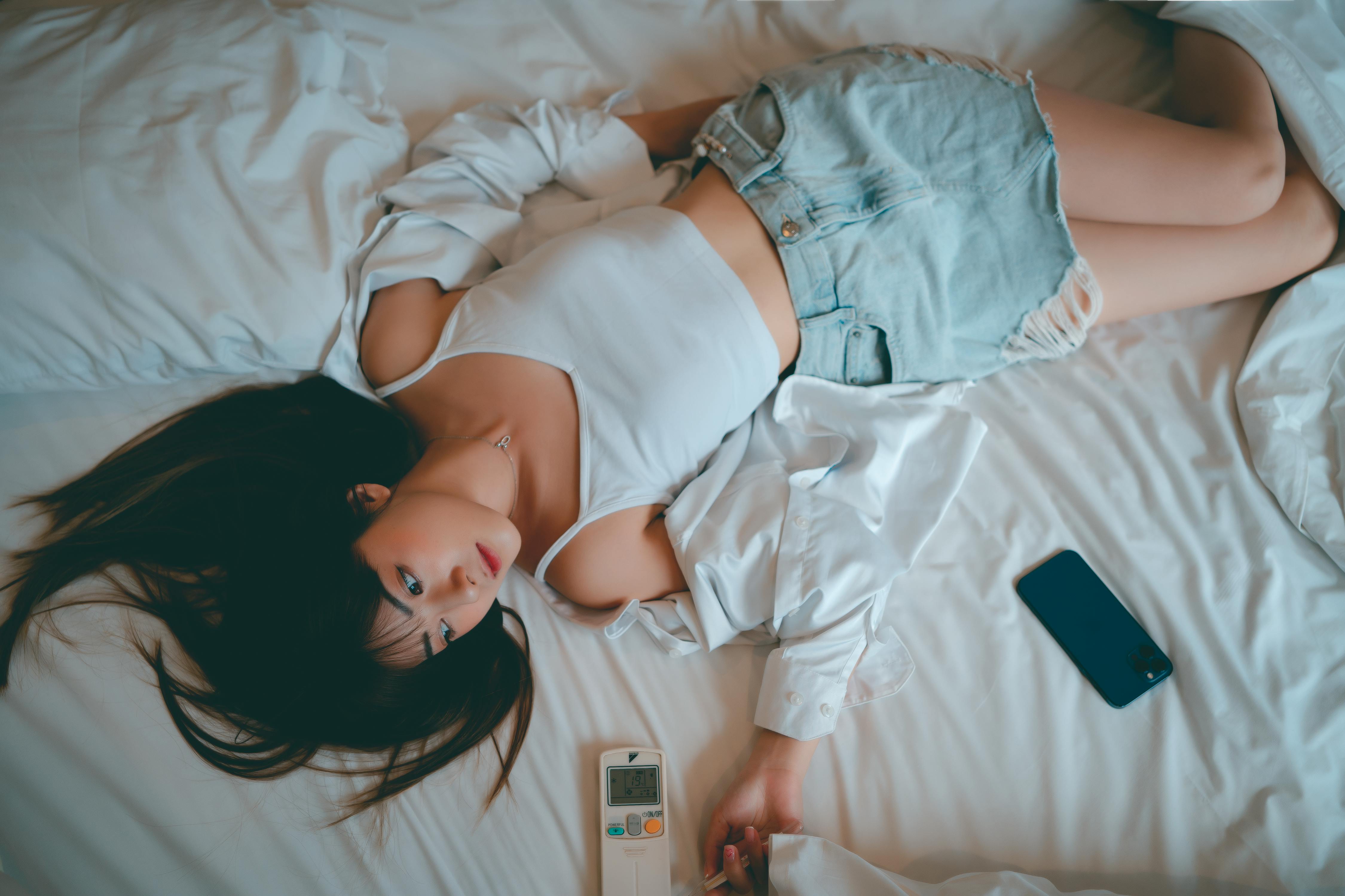 A woman in casual attire relaxes on a bed with a phone and remote control nearby.