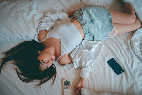 Free A woman in casual attire relaxes on a bed with a phone and remote control nearby. Stock Photo