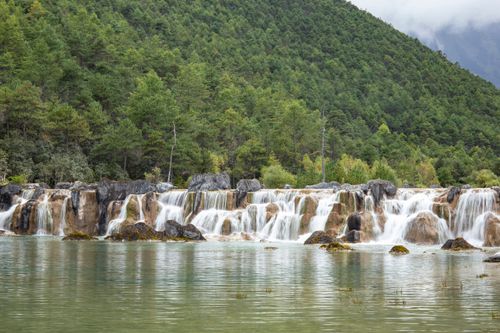 Free Breathtaking waterfall amidst lush green forests and rocky terrain in Lijiang, perfect for nature photography. Stock Photo