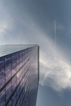 从低角度仰望摩天大楼，多云天空中有喷射轨迹反射着阳光