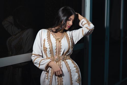 Free Sophisticated woman in an ornate dress poses gracefully by a reflective glass surface, creating a captivating portrait. Stock Photo