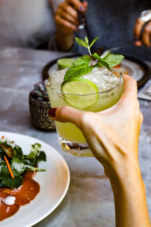 Free A hand holding a lime mint cocktail at a dining table with a salad, perfect for a refreshing drink option. Stock Photo