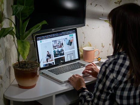 A woman works remotely at home using a laptop, seated at a cozy desk space with a potted plant and a cup.