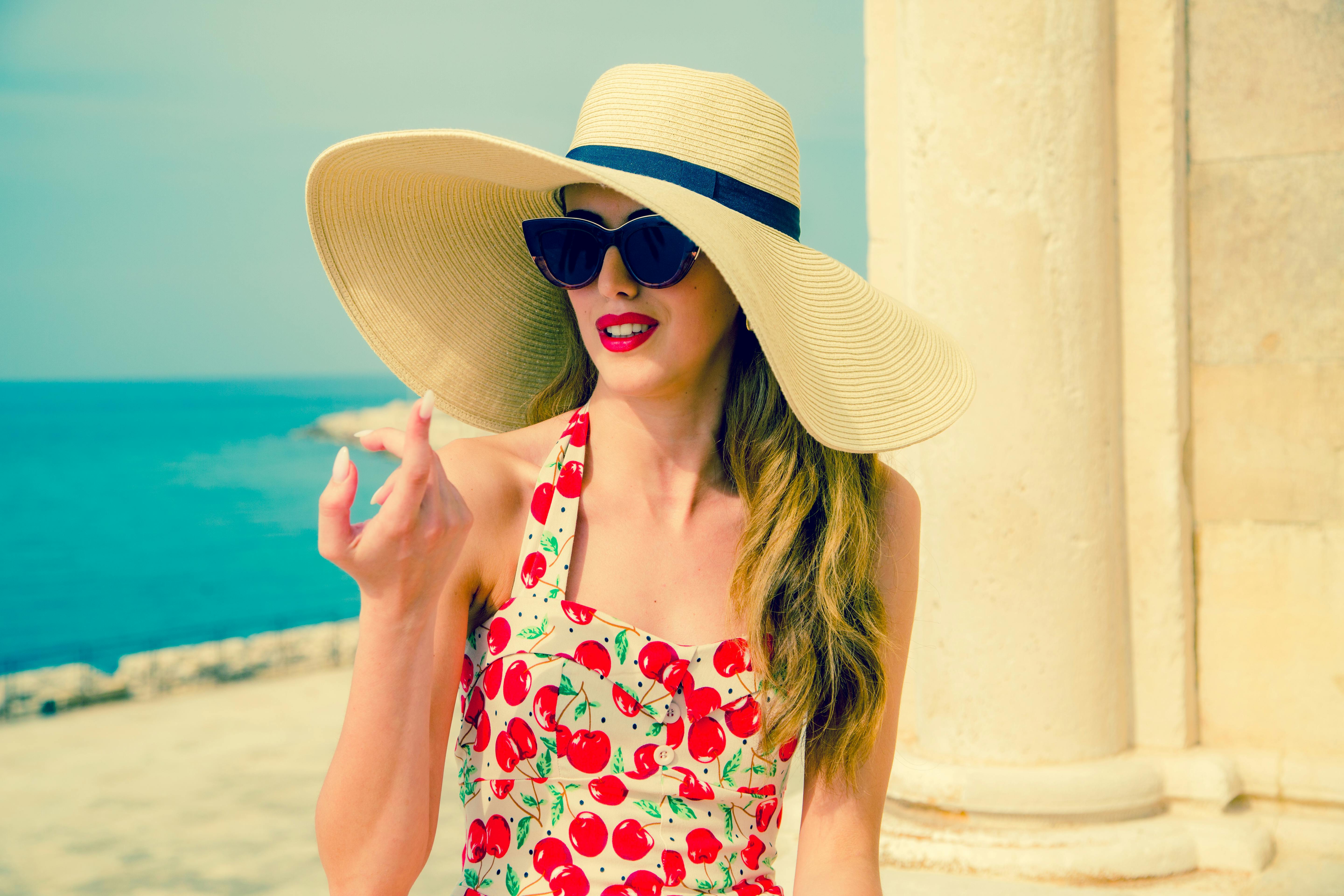 A woman with sunglasses and a sun hat enjoying a sunny day by the seaside, wearing a cherry-patterned swimsuit.