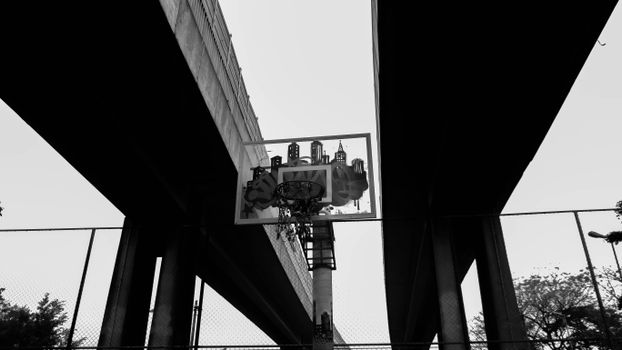 Low angle black-and-white view of a basketball hoop between two bridges, inspiring urban sports feel.