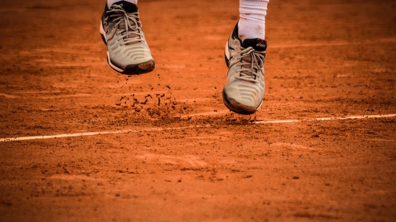 Close-up of an athlete's shoes mid-jump on a clay tennis court.