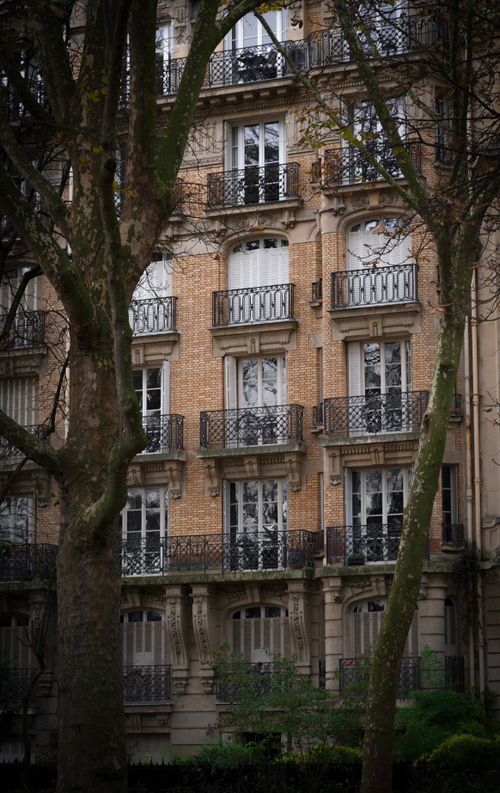 Free Classic brick building facade with intricate balconies framed by tree trunks in a city setting. Stock Photo