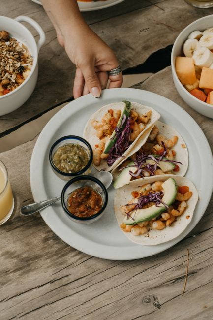 A close-up of hand holding a plate of tacos, garnished with avocado and red cabbage, served with salsas.