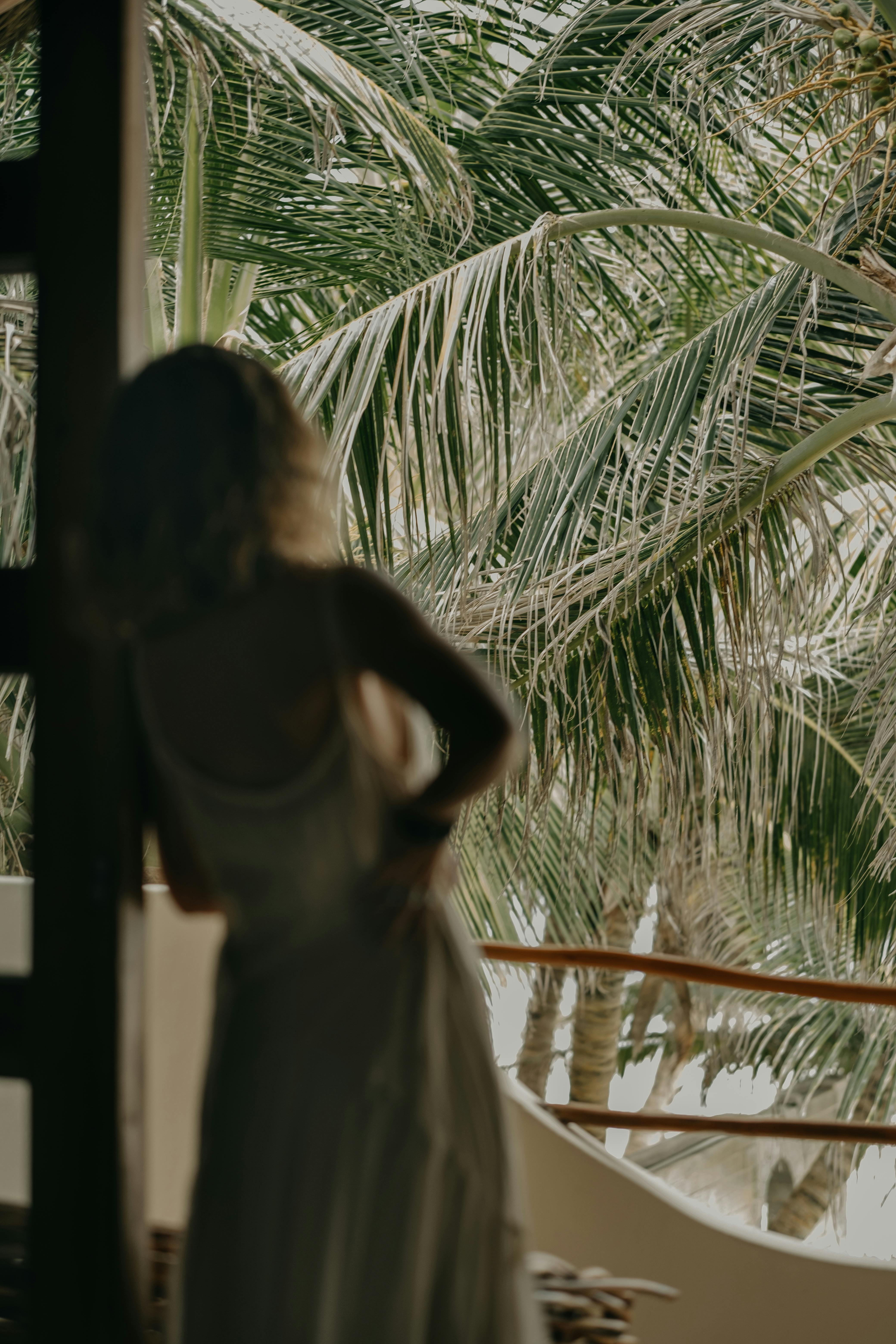Silhouette of a woman in a white dress leaning against a railing with palm trees in the background.