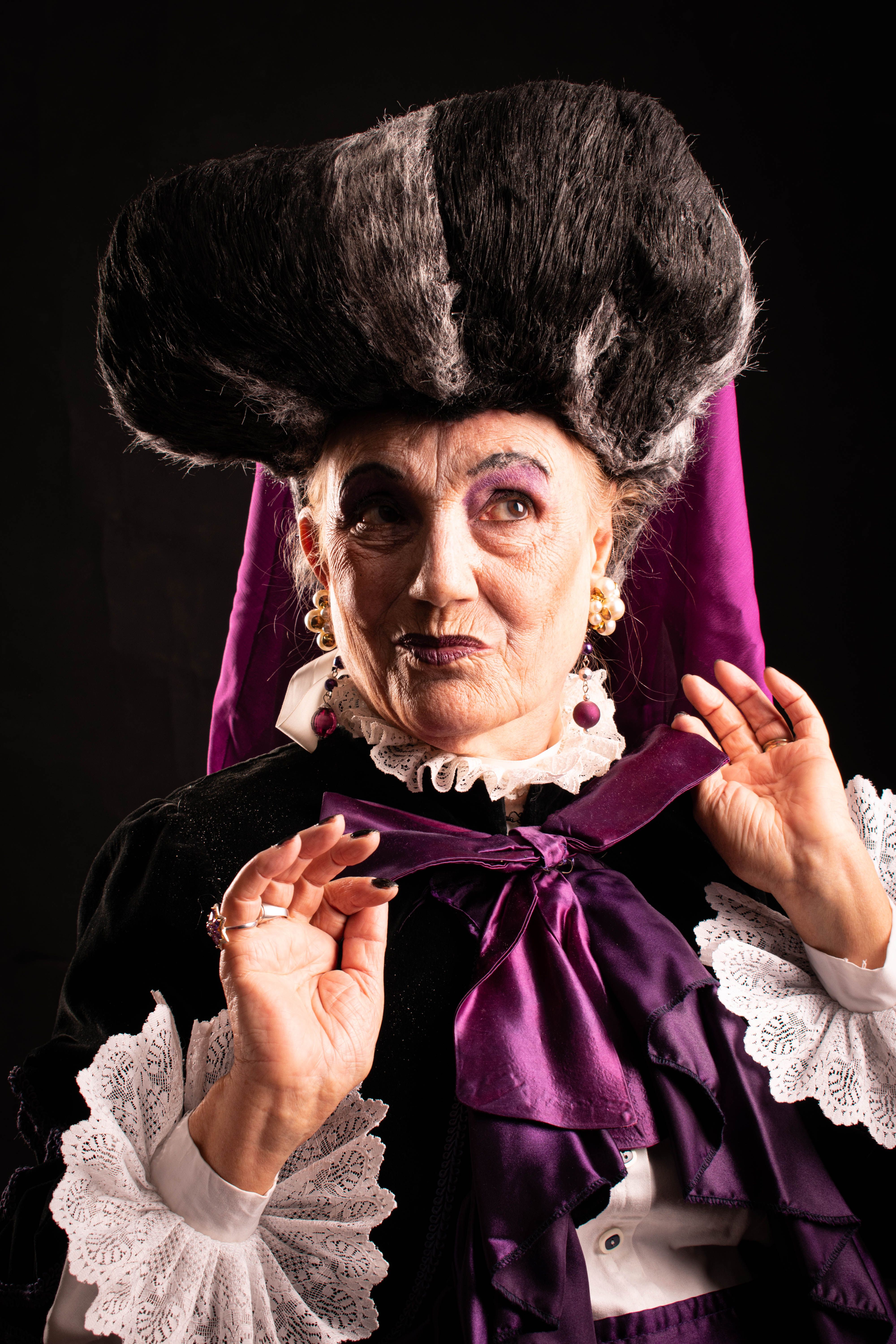 Portrait of an elderly woman in a dramatic costume with headdress against a black background.