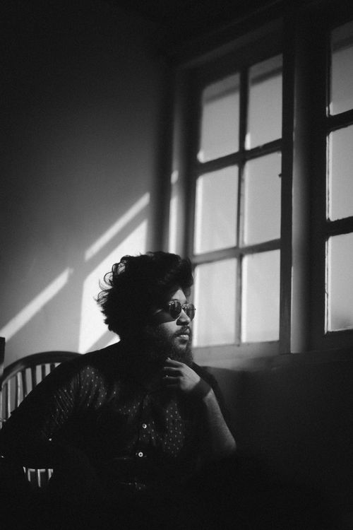 Free Contemplative man wearing sunglasses sits by a window in a shaded room. Stock Photo