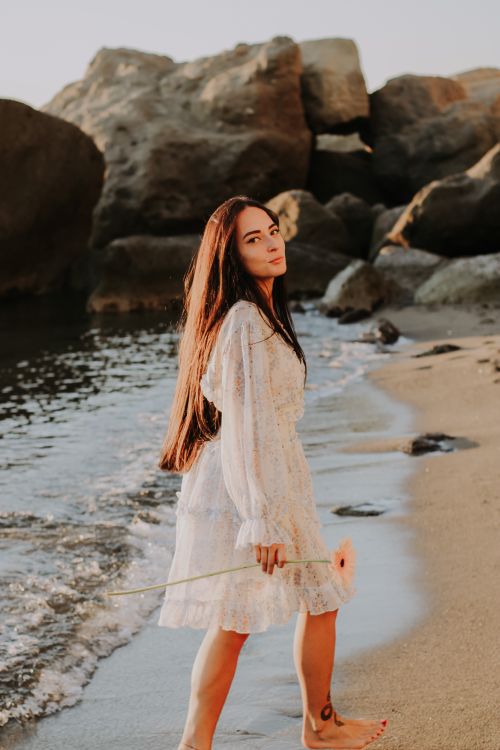 Free A woman in a white dress walks along the beach, holding a flower, at sunset. Stock Photo
