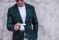 在迪拜，一位穿着绿色夹克的时尚男子靠在纹理墙上，拿着相机