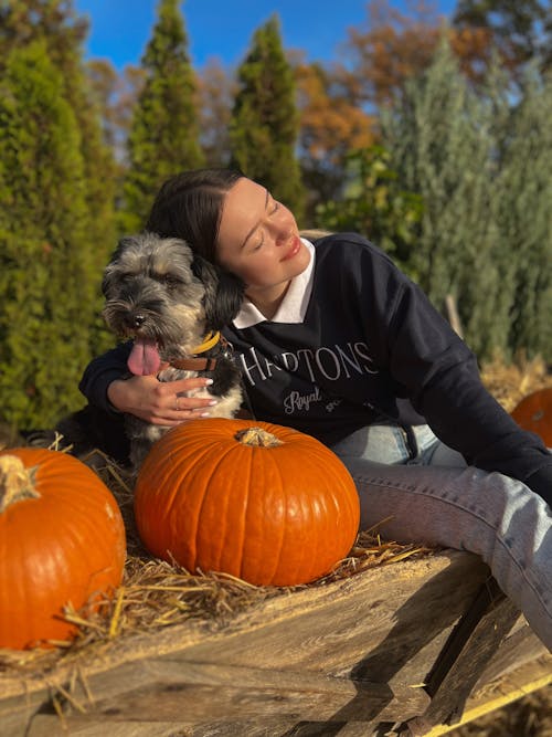 A Woman Sitting on Wooden Cart with Pumpkins Hugging a Dog
