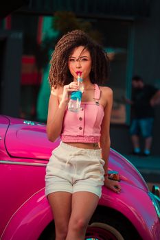 A stylish woman drinking water while leaning on a pink car in an urban setting.