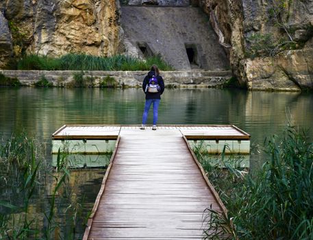 A woman stands on a wooden pier by a lake, wearing a backpack, surrounded by natural rock formations.