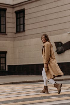 Young woman in a beige trench coat and brown boots crossing a street, showcasing urban style.